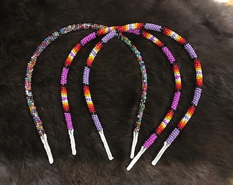 May include: Four beaded headbands in a variety of colors, including purple, red, yellow, and green. The headbands have white tips and are arranged on a dark, textured surface. These accessories are perfect for adding a pop of color.