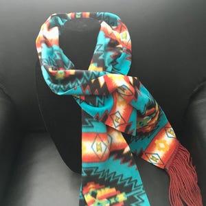 May include: A turquoise, orange, and black patterned scarf with a southwestern design. The scarf has a fringe at the ends and is displayed on a black mannequin head. The scarf is made of a soft material.