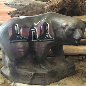 Könnte beinhalten: Eine graue Keramik-Bärenfigur mit einem bemalten Design eines Sonnenuntergangs und drei Gebäuden auf dem Rücken. Der Bär steht auf einer Holzoberfläche.