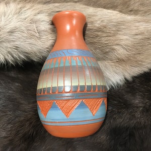 Puede incluir: Un jarrón de terracota pintado a mano con un diseño geométrico en tonos azules, marrones y naranjas. El jarrón tiene un cuerpo redondeado y un cuello estrecho.