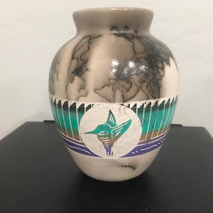 May include: A rounded ceramic vase with a smooth, light-colored finish featuring dark, wispy smoke-like markings. A horizontal band wraps around the center, displaying teal, brown, and purple stripes with a white circular medallion showing a hummingbird.