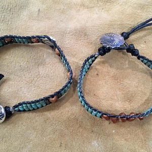Peut inclure: Deux bracelets composés d'un cordon noir tressé avec de petites perles turquoise et des perles marron intercalées. Chaque bracelet est fermé par un bouton en métal argenté de forme ronde orné d'un motif géométrique en relief.