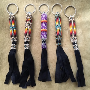 May include: Five beaded keychains with silver rings and black suede tassels. The beaded sections feature colorful patterns in red, orange, yellow, blue, and white. Each keychain has a unique design, showcasing intricate beadwork.