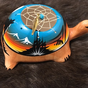 May include: A hand-painted ceramic turtle with a dreamcatcher inside. The turtle is painted with a sunset scene and mountains. The dreamcatcher has a turquoise bead and a gold feather.