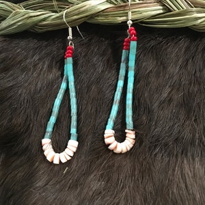 May include: Handmade turquoise and red beaded earrings with a loop of white shell beads at the bottom. The earrings hang from silver-colored hooks. The background is a dark fur.