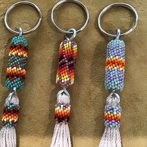 May include: Three beaded keychains with tassels. Each keychain features a cylindrical bead with a colorful geometric pattern and a long, white tassel. The beads are turquoise, brown, and purple.