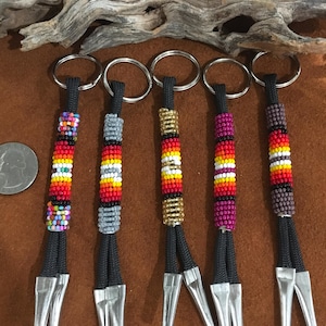 Op de afbeelding: Vijf met kralen versierde sleutelhangers met zilveren ringen en zwarte koorden. De sleutelhangers hebben kleurrijk kralenwerk in rode, gele, oranje en zwarte patronen. Een munt ligt naast de sleutelhangers ter vergelijking van de grootte.