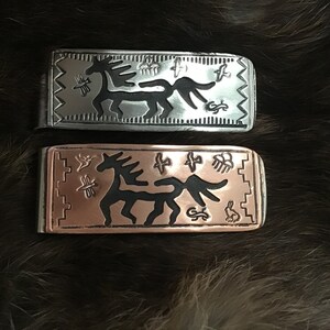Könnte beinhalten: Zwei Geldklammern aus Metall mit einem gravierten Pferdemotiv. Eine Klammer ist silberfarben, die andere kupferfarben. Beide zeigen ein stilisiertes Pferd, Vögel und geometrische Muster und bieten eine rustikale, künstlerische Ästhetik.