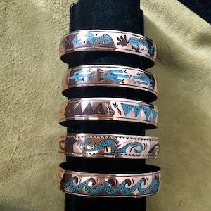 May include: A stack of copper-colored metal bracelets with turquoise inlay. Each bracelet features a different design, including geometric shapes, waves, and desert-themed motifs. The bracelets are displayed on a black stand.