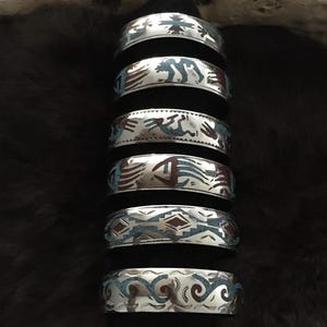 Op de afbeelding: Zeven zilverkleurige manchetarmbanden met ingelegde turquoise en bruine ontwerpen. Elke armband heeft een uniek patroon, waaronder geometrische vormen en diermotieven. De armbanden zijn gestapeld, met een donkere achtergrond. De ontwerpen doen denken aan Indiaanse kunst.