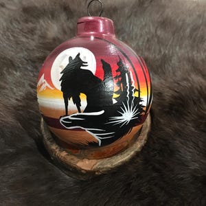 May include: Hand-painted Christmas ornament featuring a wolf howling at the moon. The ornament has a burgundy top and a landscape scene with a full moon, mountains, and trees. The wolf is silhouetted against a sunset sky.