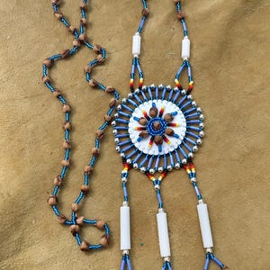 Op de afbeelding: Een kralenketting met een cirkelvormig medaillon met een wit midden, blauwe en zilveren spaken en accenten in rood, geel en zwart. De ketting bestaat uit blauwe kraaltjes afgewisseld met bruine natuurlijke zaden en drie witte buisvormige kralen.