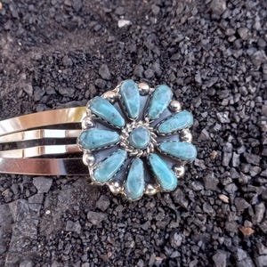 May include: A silver-toned metal hair clip featuring a circular floral ornament. The ornament is composed of twelve teardrop-shaped turquoise stones arranged in a petal pattern around a central round turquoise stone, all set in a silver frame.