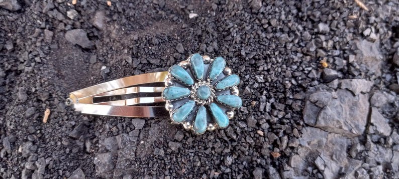 May include: A silver-toned metal hair clip featuring a circular floral ornament. The ornament is composed of twelve teardrop-shaped turquoise stones arranged in a petal pattern around a central round turquoise stone, all set in a silver frame.