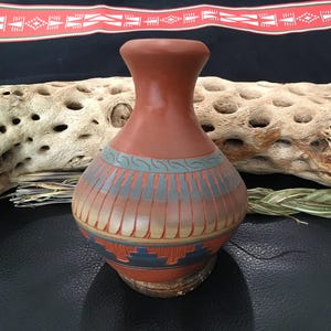 May include: A small, hand-crafted pottery vase with a rounded body and a narrow neck. The vase is decorated with a geometric pattern in shades of blue, green, and red.