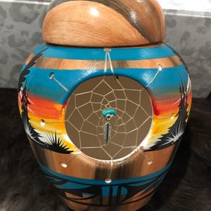 May include: A ceramic jar with a dreamcatcher design. The jar is painted with a sunset scene and has a hole in the side to allow the dreamcatcher to hang through. The dreamcatcher is made of beads and feathers.