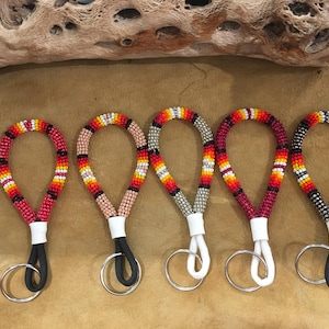May include: Five beaded keychains in red, black, white, yellow, and orange. Each keychain has a loop of beads attached to a white base and a black or white cord with a silver keyring. The keychains are displayed on a brown surface.