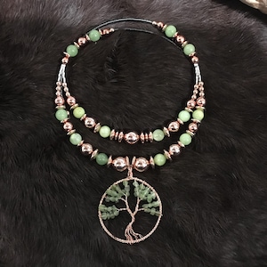 Könnte beinhalten: Eine mehrschichtige Halskette mit grünen Perlen und kupferfarbenen Metallakzenten. Die Halskette hat einen Anhänger mit einem Drahtbaum-Design. Der Schmuck wird auf einer dunklen, strukturierten Oberfläche präsentiert.