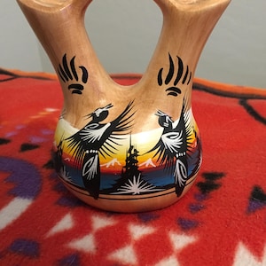 Puede incluir: Un jarrón de color marrón claro con dos picos y un asa. El jarrón presenta una escena pintada de pájaros en vuelo contra un fondo de puesta de sol. Huellas de patas de oso negras están pintadas cerca del asa. El jarrón es probablemente de cerámica.