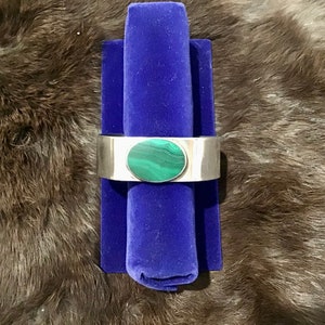 May include: A silver cuff bracelet with a round green malachite stone set in the centre. The bracelet is displayed on a blue velvet cushion.