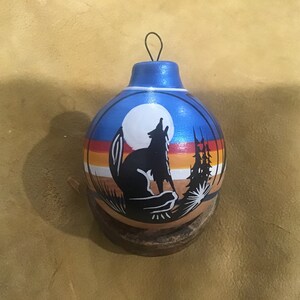May include: A blue and brown hand-painted Christmas ornament with a silhouette of a wolf howling at the moon. The ornament has a white moon, a red and yellow sunset, and a brown forest in the background.