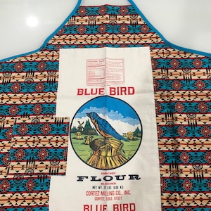 May include: A blue and white kitchen apron with a Native American inspired pattern. The apron has a large pocket with a vintage Blue Bird Flour sack sewn into it. The flour sack features a bluebird perched on a stalk of wheat. The text on the flour sack reads "Enriched Bleached Flour, Net Wt. 20 lbs. 9.08 kg, Cortez Milling Co., Inc., Cortez, Colo. 81321, Blue Bird."