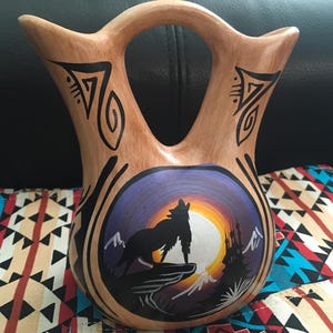 Pode incluir: Vaso de cerâmica pintado à mão com base castanha e desenhos tribais pretos. O vaso apresenta uma imagem central de um lobo uivando contra um pôr do sol. O vaso tem uma alça e uma forma única.