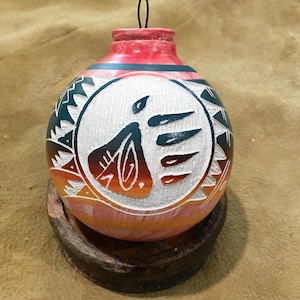 May include: Hand-painted, round, decorative ornament with a tribal design. The ornament features a red top, a white center with a stylized figure, and orange and teal accents. It sits on a small, round wooden base.