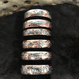 May include: A stack of six copper-colored metal bracelets with etched designs. The bracelets feature various Southwestern-inspired motifs, including horses, cacti, and geometric patterns. The bracelets are displayed against a dark, textured background.