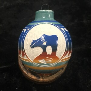 Puede incluir: Adorno de cerámica pintado a mano con una parte superior turquesa y un diseño circular con una silueta azul de un oso. El adorno tiene un fondo blanco con detalles en azul, naranja y turquesa, y se asienta sobre una base de madera.