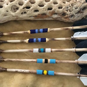 May include: Four handcrafted arrows with stone arrowheads and feather fletching. The wooden shafts are decorated with colorful beaded bands in blue, white, and yellow. The arrows are arranged on a tan surface.