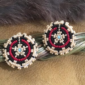 Può includere: Un paio di orecchini a bottone con perline. Ogni orecchino presenta un design circolare con un anello esterno color crema, cerchi concentrici neri e rossi e un centro azzurro. Gli orecchini sono esposti su una superficie color cuoio.