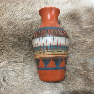 Puede incluir: Un pequeño jarrón de terracota con un diseño geométrico en tonos azules, marrones, blancos y amarillos. El jarrón está decorado con un patrón de inspiración suroeste.