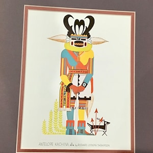 Puede incluir: Una ilustración colorida de un Kachina Antílope, una figura tradicional de espíritu Hopi, con un traje estampado en amarillo, azul, rojo y blanco. El Kachina sostiene un bastón y tiene un perro blanco y negro a sus pies. La imagen está titulada "ANTELOPE KACHINA 1974 by RICHARD JOSEPH THOMPSON".