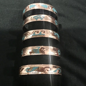 May include: Six copper-coloured metal bracelets with turquoise and brown inlay designs. Each bracelet features a different pattern of geometric shapes and stylized figures. The bracelets are stacked vertically on a black display stand.