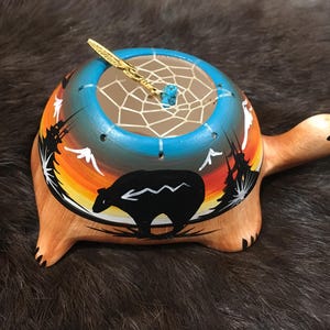May include: A hand-painted wooden turtle with a dreamcatcher inside. The turtle is decorated with a sunset scene, a bear, and trees. The dreamcatcher is made of brown leather and has a turquoise stone in the center.