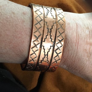 May include: A copper cuff bracelet with an etched geometric design. The bracelet features a series of parallel bands with repeating diamond and stepped patterns. The cuff is worn on a wrist, showcasing the intricate metalwork and warm tones of the copper.