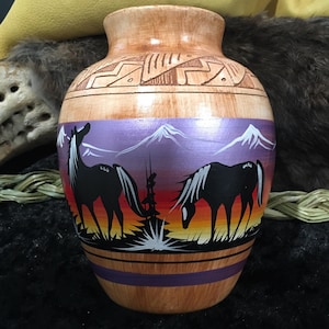 Pode incluir: Um vaso de cerâmica pintado à mão com um design do sudoeste. O vaso apresenta uma base castanha clara com uma cena de pôr do sol roxa e laranja retratando dois cavalos pretos, montanhas e um padrão geométrico decorativo ao redor do pescoço.
