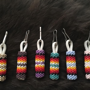 May include: Seven beaded zipper pulls with a variety of colors, including red, yellow, blue, green, purple, black, and white. The zipper pulls are attached to white cords and have silver metal clips.