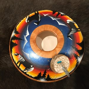 Pode incluir: Tigela de madeira pintada à mão com uma abertura central, apresentando um design vibrante de silhuetas pretas, árvores e pássaros contra um gradiente de pôr do sol de laranja, amarelo e azul. Um pequeno apanhador de sonhos com uma conta turquesa e um pingente de pena está anexado.