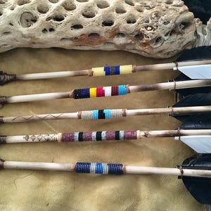 May include: Five handcrafted arrows with wooden shafts, stone arrowheads, and feather fletching. The shafts are adorned with colorful beaded bands in various patterns. The arrows rest on a tan surface.