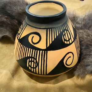 May include: A hand-painted ceramic vase with a rounded body and a narrow neck. The vase features a tan background with black geometric patterns, including triangles, lines, and spiral designs. The rim is black.