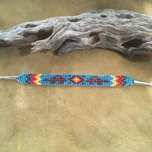Puede incluir: Una pulsera de cuentas con fondo turquesa y patrones geométricos en rojo, naranja y marrón. La pulsera tiene extremos de metal plateado y un cierre. El diseño recuerda al arte nativo americano.