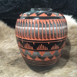 Puede incluir: Un jarrón de cerámica hecho a mano con cuerpo redondeado y cuello estrecho. El jarrón presenta un patrón geométrico negro, naranja y gris. El diseño incluye líneas verticales y formas triangulares. Es un artículo decorativo.