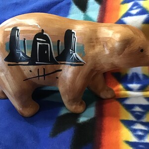 Puede incluir: Una figurita de oso de madera pintada con un diseño de ciudad en blanco y negro. El oso es marrón y tiene un glaseado marrón claro. El diseño de la ciudad incluye un edificio con una puerta y una ventana.