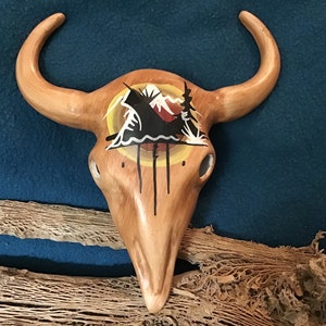 May include: A wooden bull skull with a painted design of a mountain scene with a yellow sun and black trees. The skull is painted in shades of brown, yellow, black, and white.