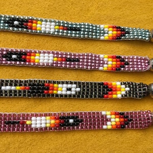 Puede incluir: Primer plano de cuatro pulseras de cuentas con un diseño nativo americano. Las pulseras presentan una variedad de colores, incluyendo negro, blanco, rojo, amarillo y morado. Cada pulsera tiene una cuenta de color plateado en el extremo.
