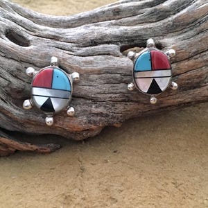 May include: A pair of silver-toned turtle-shaped earrings. Each earring features a mosaic design with red, turquoise, black, white, and gray segments. The earrings are displayed on a piece of driftwood against a sandy background.