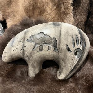 May include: A carved wooden bear figurine in shades of grey and black. The bear has a sun and paw print design. The figurine is resting on a brown fur surface.