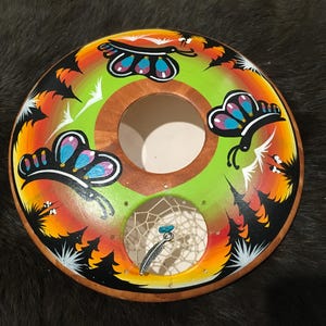 Pode incluir: Um apanhador de sonhos de madeira com um design pintado em verde, laranja e preto com borboletas. O apanhador de sonhos tem uma pequena abertura redonda no centro com uma pena branca e uma conta azul.